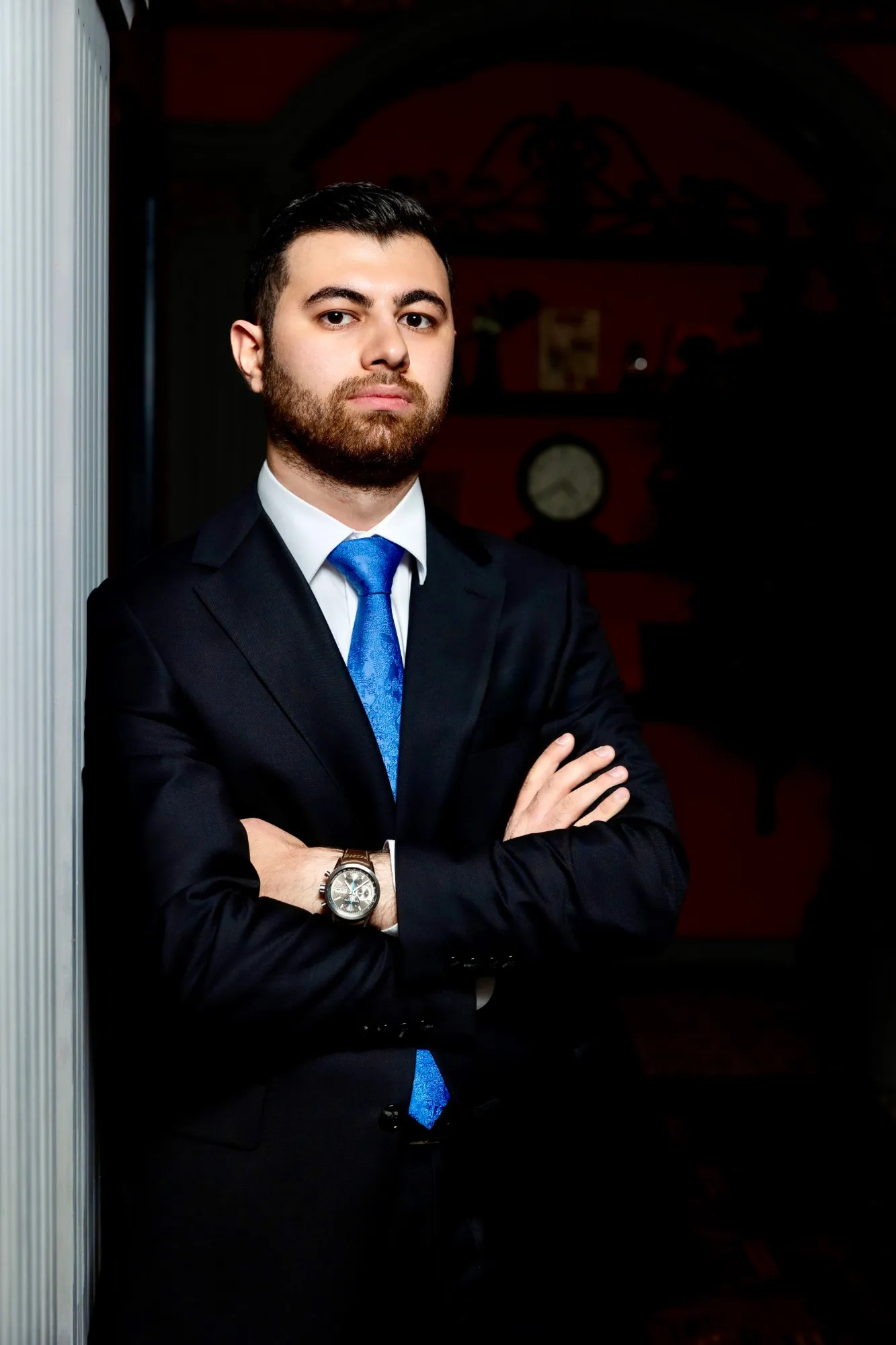 Riad Ibadulla in a dark suit and blue tie, arms folded, standing beside a white column.